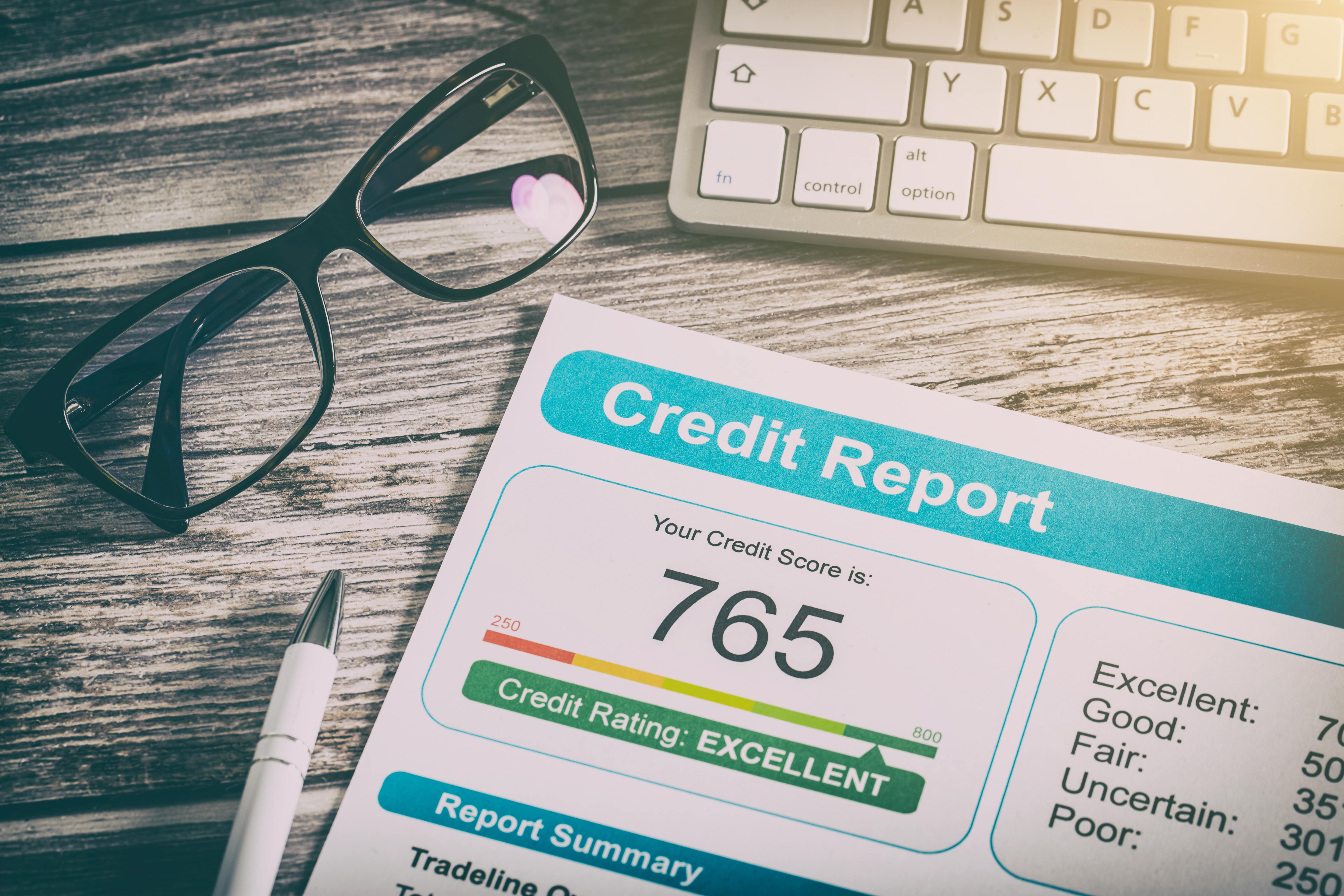 Close-up of a credit report showing an excellent credit score of 765, with glasses and a keyboard in the background.