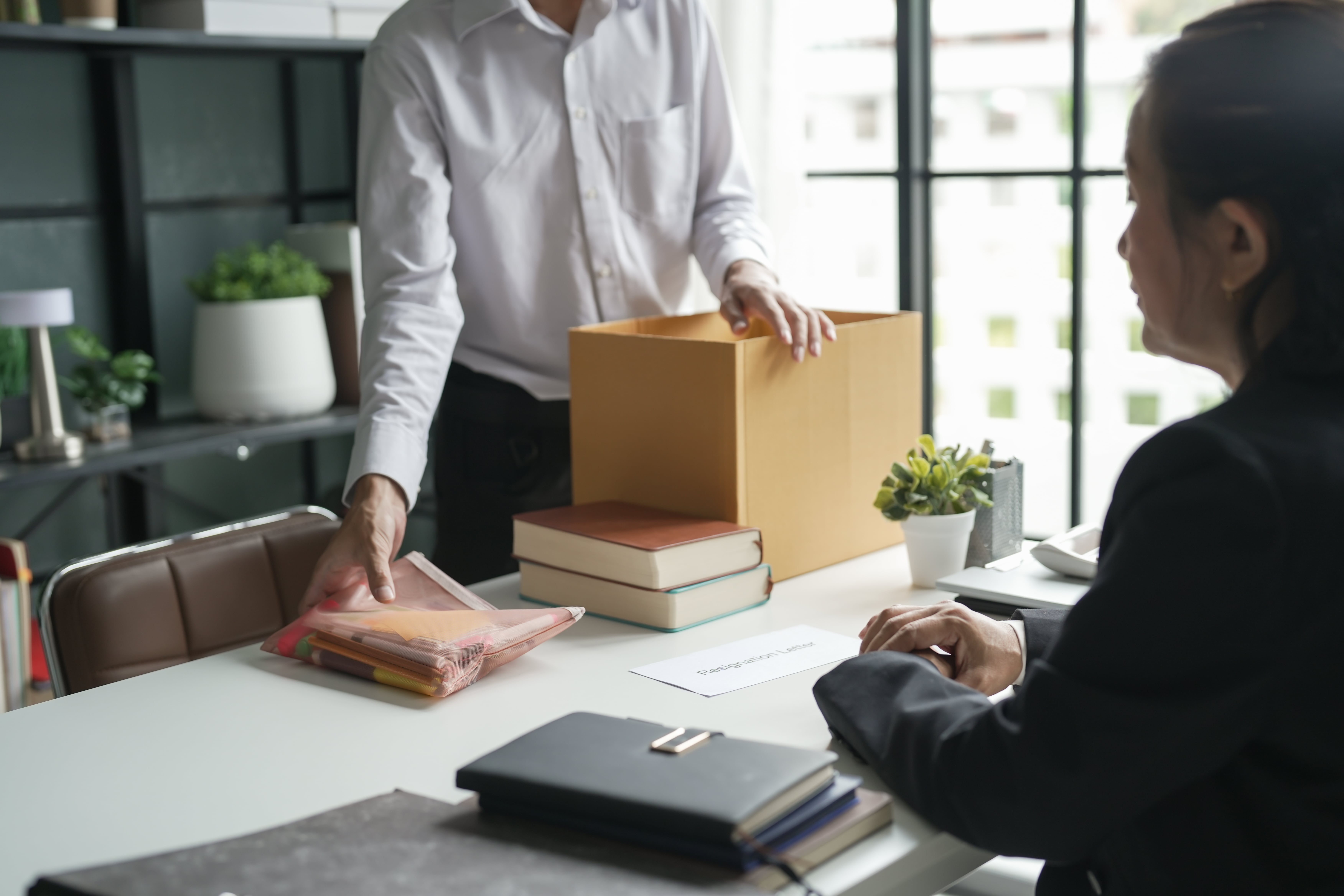 A professional packing office supplies into a cardboard box while another person watches, symbolizing the termination or resignation of a property manager.