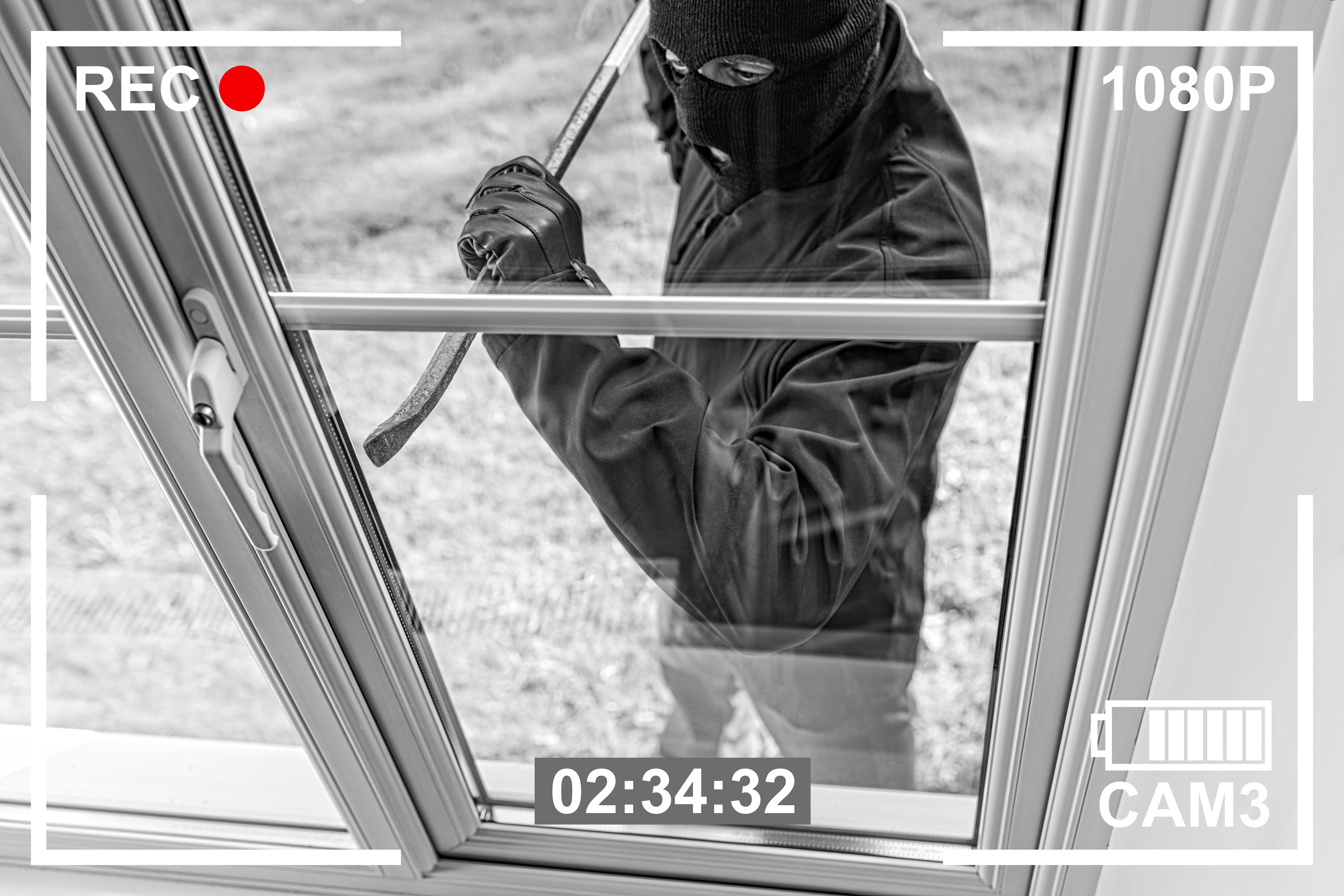 A burglar in a ski mask attempting to break into a house through a window — captured on a security camera. This image is ideal for discussing home security, crime prevention, or vacant home watch services.