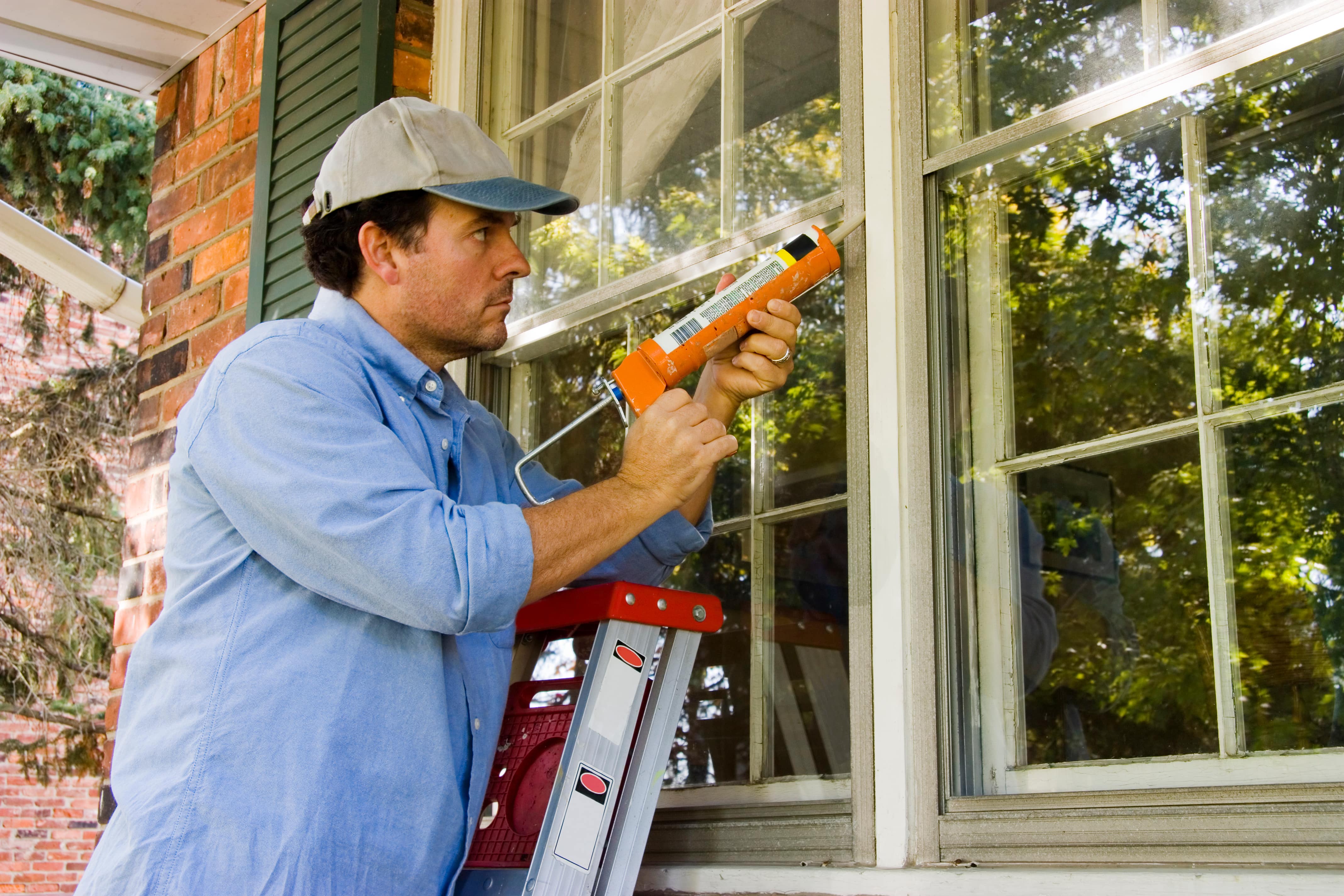 Man in a cap using a caulking gun to seal a window frame while standing on a step ladder outside a brick house.
