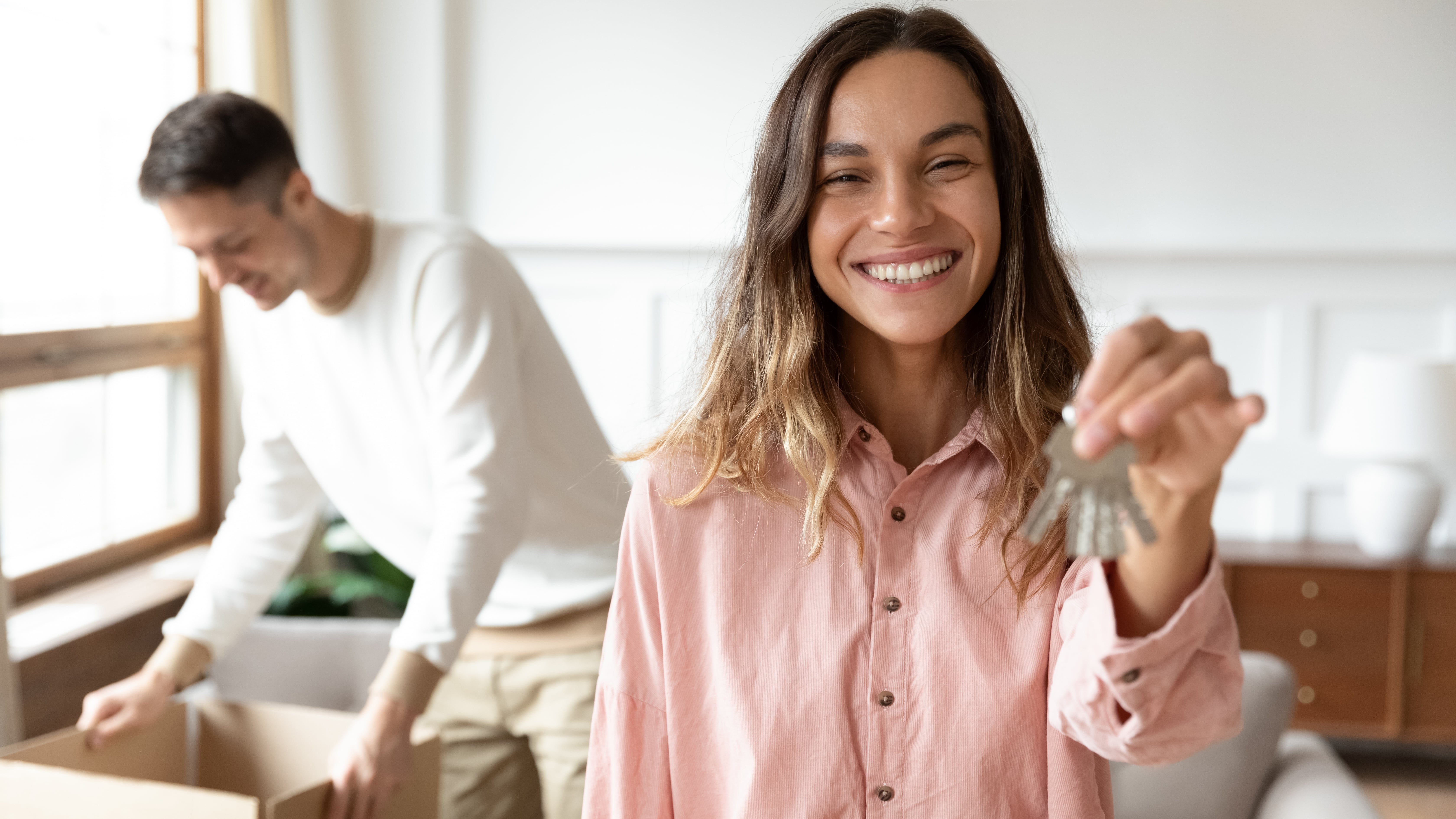 Happy woman holding a set of house keys with a man unpacking boxes in the background, symbolizing a successful move-in experience.