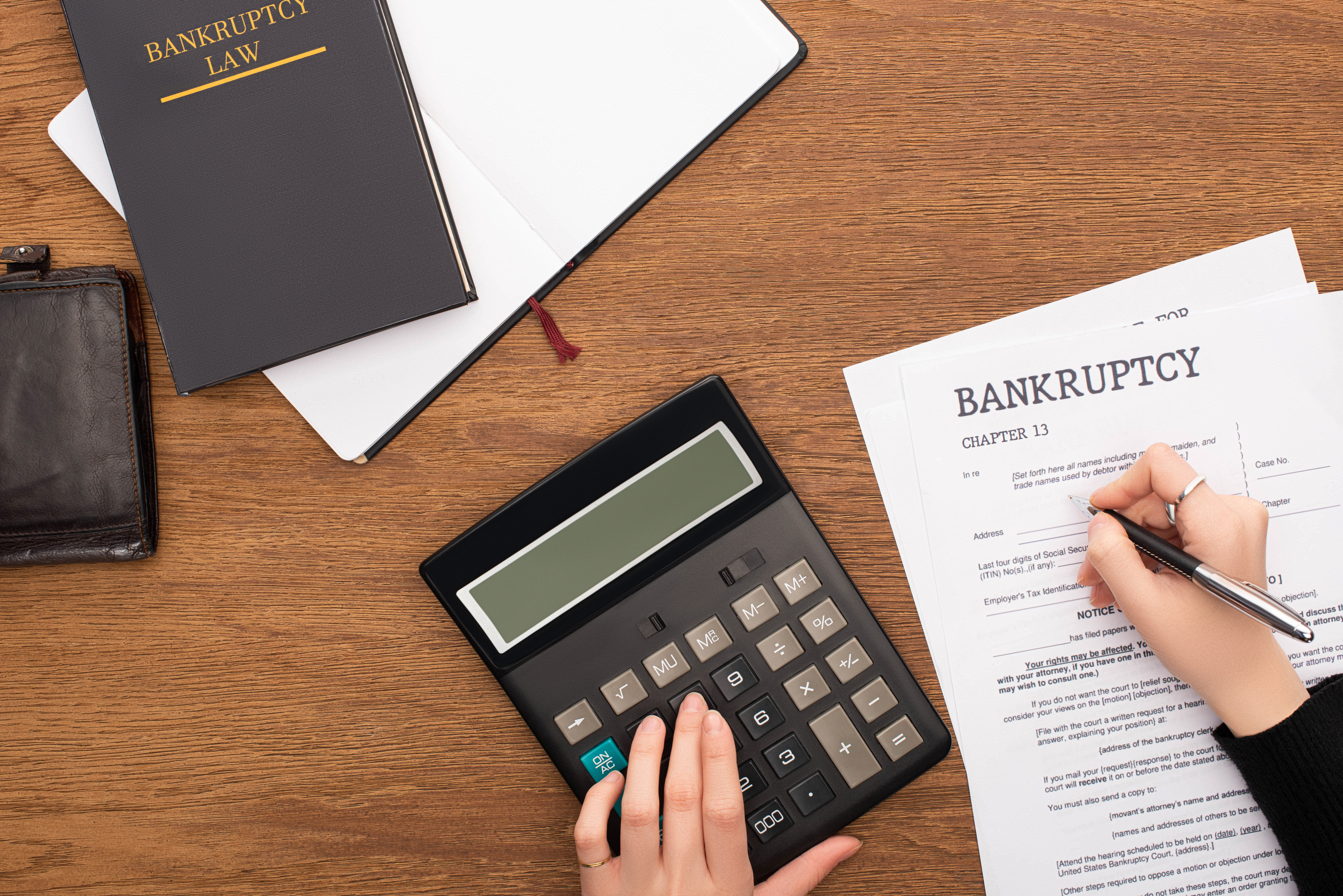 Top-down view of a person filling out a Chapter 13 bankruptcy form at a wooden desk, with a black calculator, an open notebook, a leather wallet, and a hardcover book titled ‘Bankruptcy Law’ nearby.