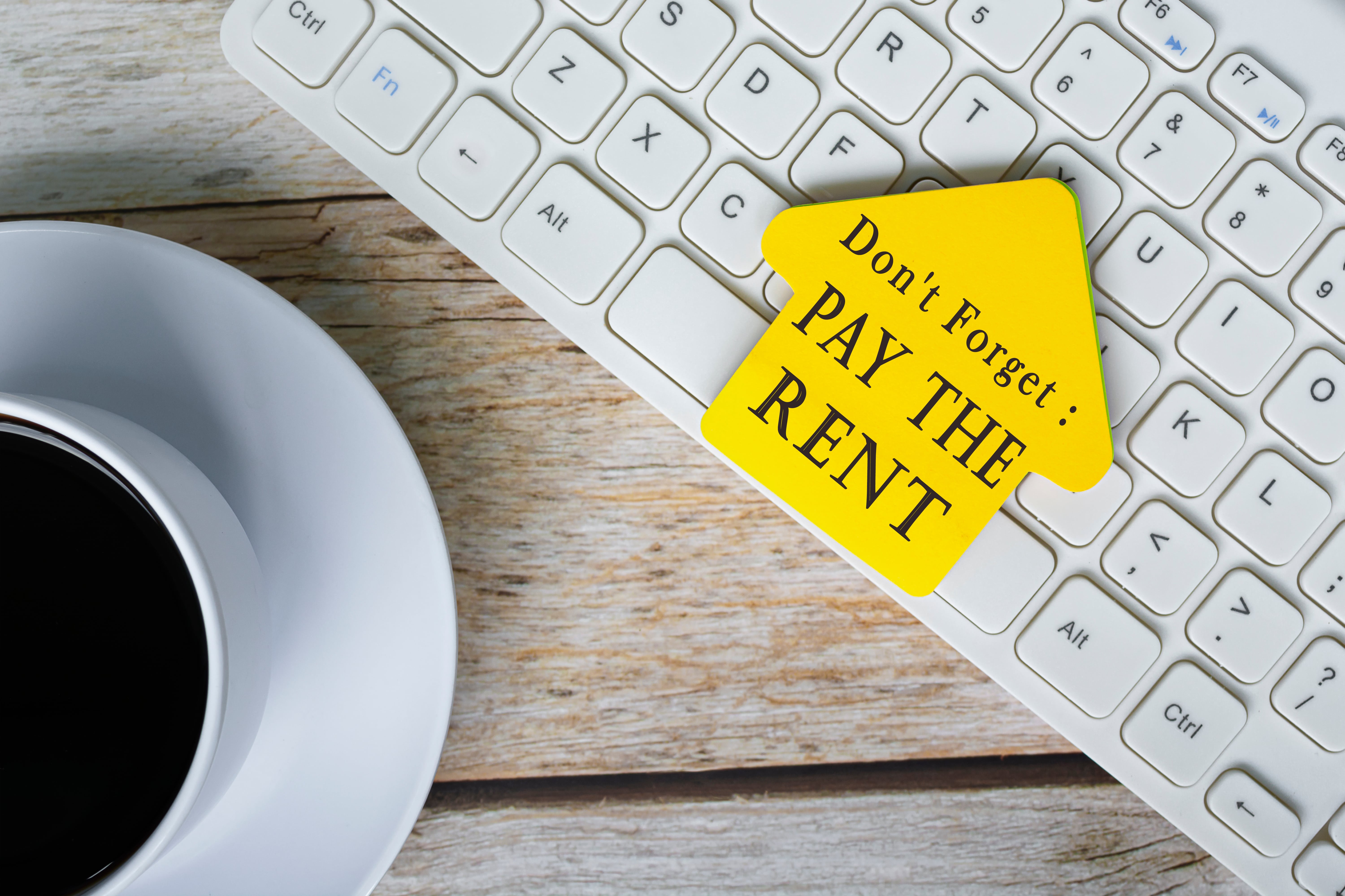 Yellow house-shaped sticky note on a keyboard that reads ‘Don’t Forget: PAY THE RENT,’ next to a cup of coffee.
