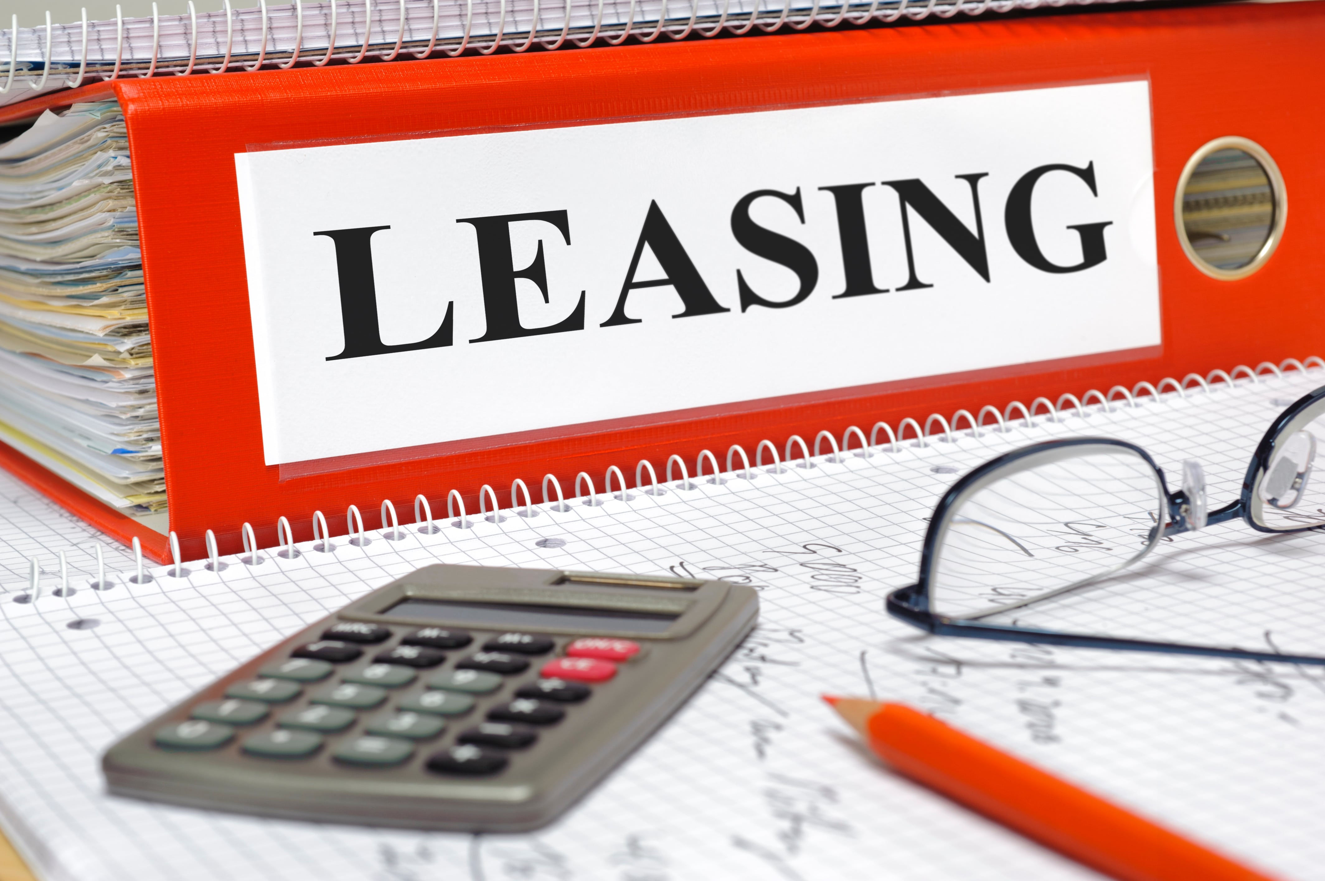 Large red binder labeled “LEASING” on a desk with a calculator, eyeglasses, a pencil, and graph paper covered in financial notes.