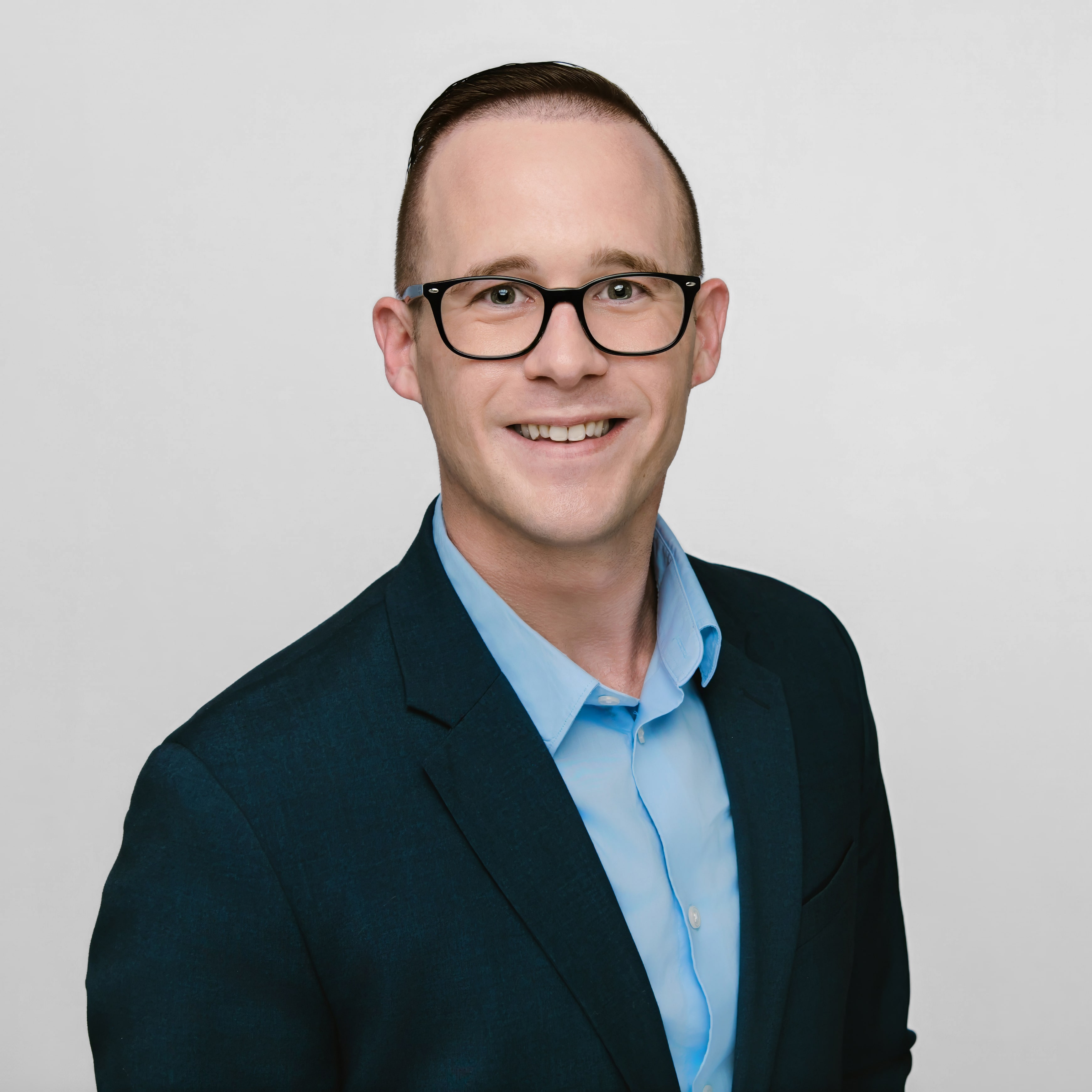 Professional headshot of Alex Zweydoff, wearing a dark blazer and light blue button-up shirt, smiling confidently against a plain light-gray background. He has short brown hair, glasses, and a clean, polished appearance.