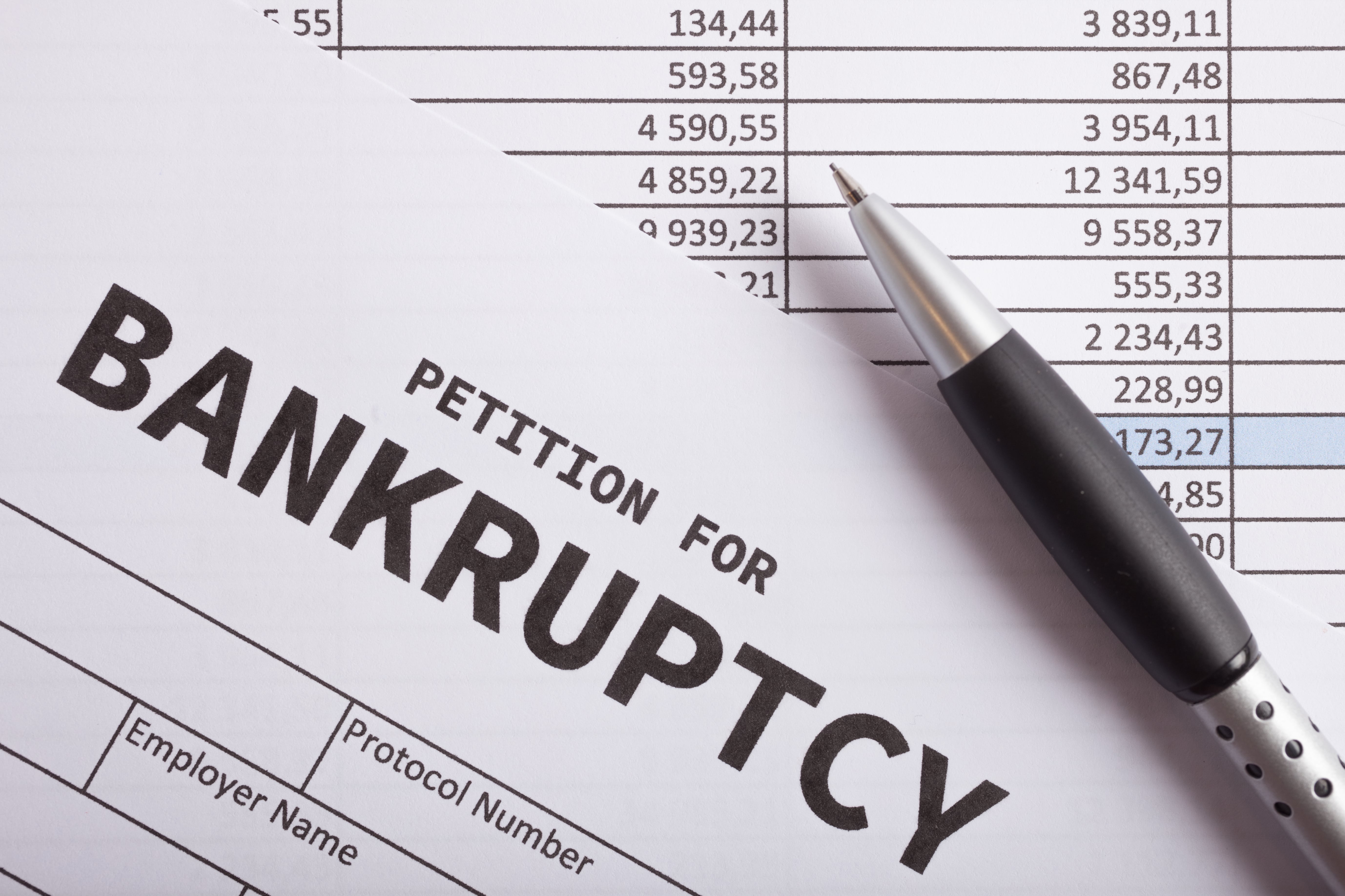 Close-up of a ‘Petition for Bankruptcy’ form placed over a financial spreadsheet with a silver and black pen resting on top, highlighting a list of monetary figures.