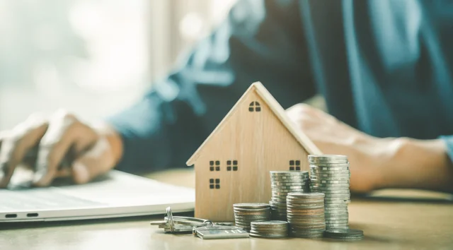 Person working on a laptop beside a small wooden house model and stacked coins on a desk, representing property investment, rental income, and financial management.