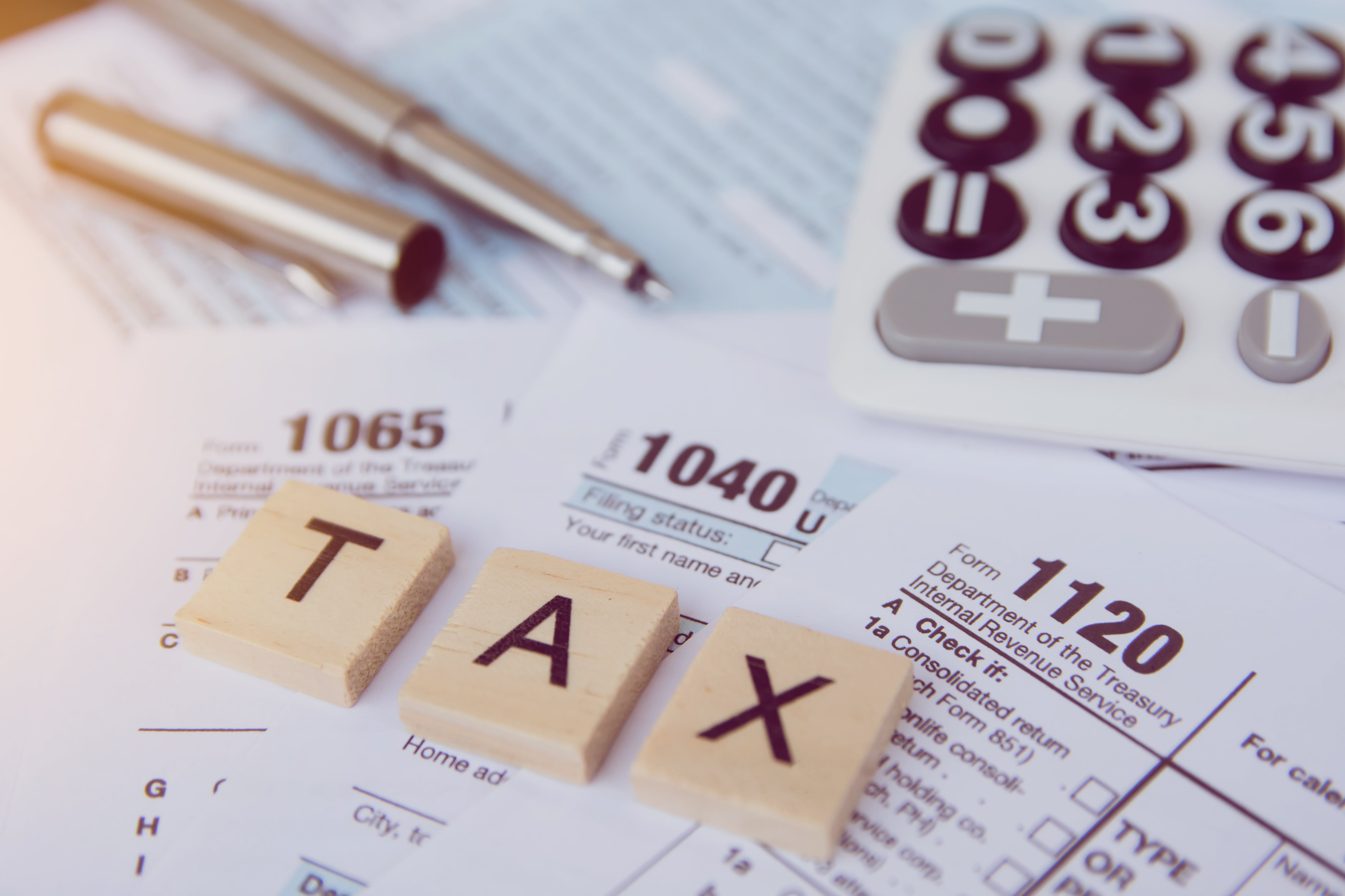 U.S. tax forms 1040, 1065, and 1120 with a calculator and ‘TAX’ spelled in wooden blocks, representing real estate tax strategies.